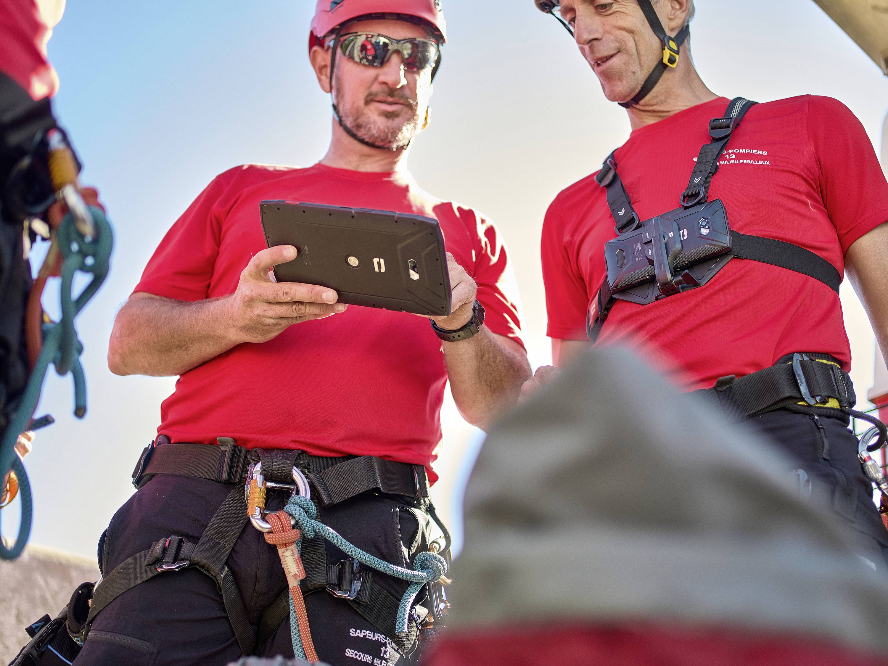 Das Bild zeigt zwei Männer bei der Nutzung eines Industrie-/Outdoor-Tablets unter freiem Himmel. Beide tragen Schutzausrüstung und Schutzhelme. Der linke Mann trägt eine Sonnenbrille und hält das widerstandsfähige Tablet, während der rechte Mann einen Brustgurt trägt und lächelt. Beide betrachten den Bildschirm des Tablets.