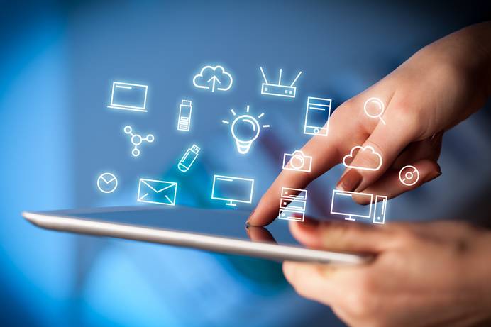 Das Bild zeigt eine Hand, welche mit einem horizontal gehaltenen Bildschirm eines Tablets interagiert. Über dem Tablet schweben weiße, leuchtende Liniensymbole auf blauem Hintergrund, die verschiedene technische Geräte darstellen, darunter einen Laptop, eine Cloud, eine Glühbirne, ein Telefon, einen Router und einen Monitor. Der Finger der Person berührt den Tablet-Bildschirm in der Nähe dieser Symbole und betont die Konnektivität im Bereich des Smart Home.