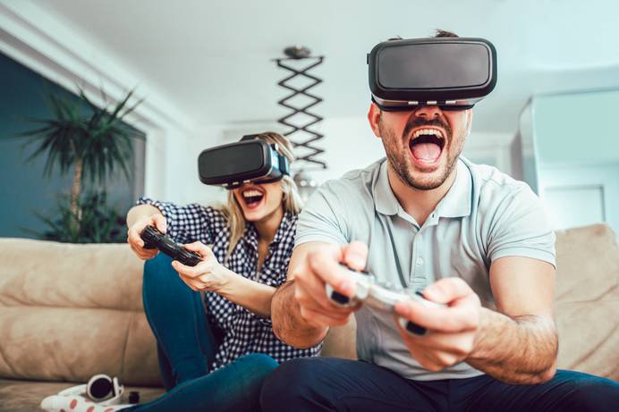 Ein Mann und eine Frau spielen lachend VR-Gaming mit Headsets und Controllern auf einer Couch.