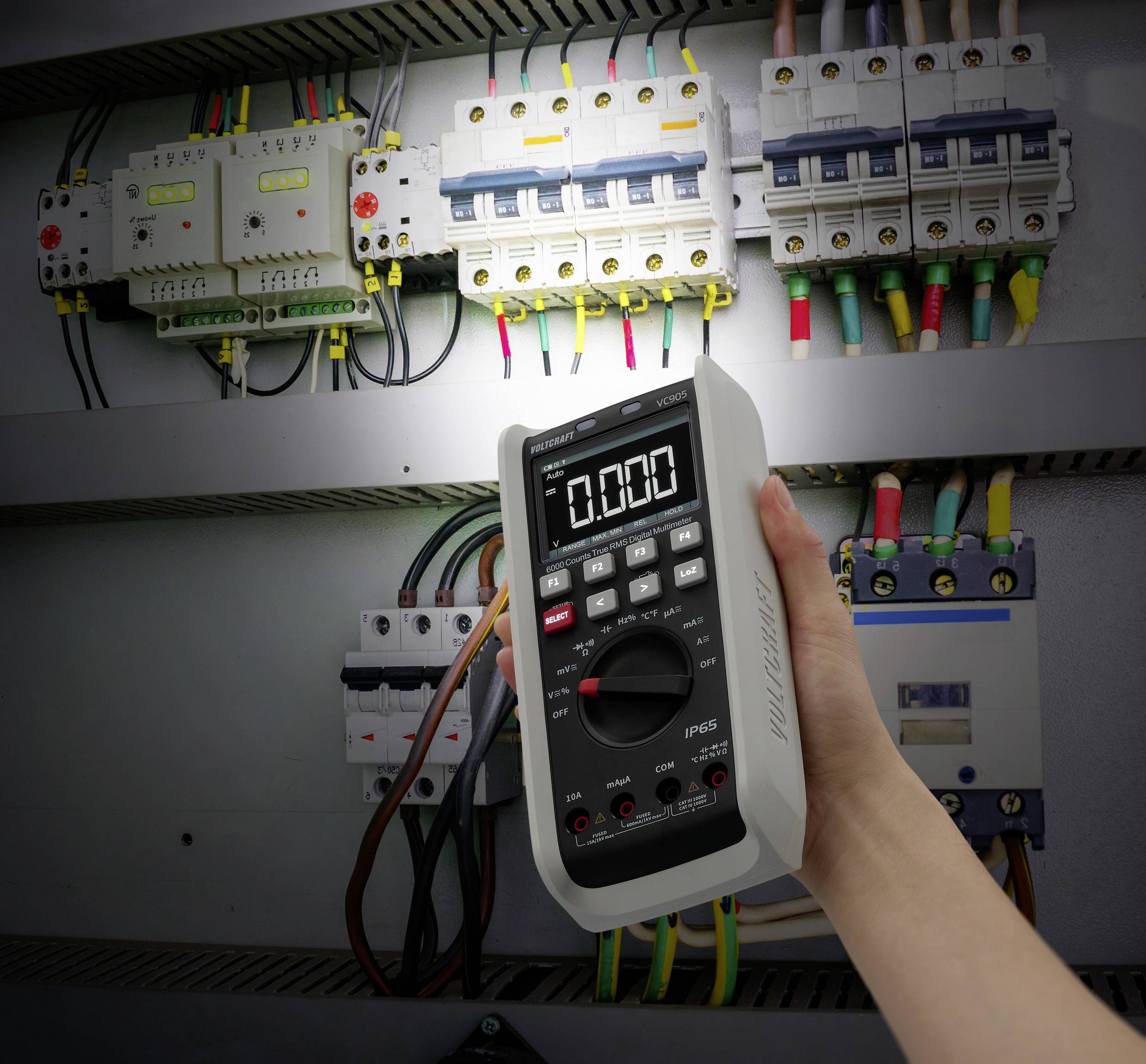 Ansicht eines geöffneten Schaltschranks. Zu sehen sind verschiedene Leitungsschutzschalter mit abgehenden Kabeln. Davor hält eine Hand ein Multimeter mit digitaler Anzeige hoch.