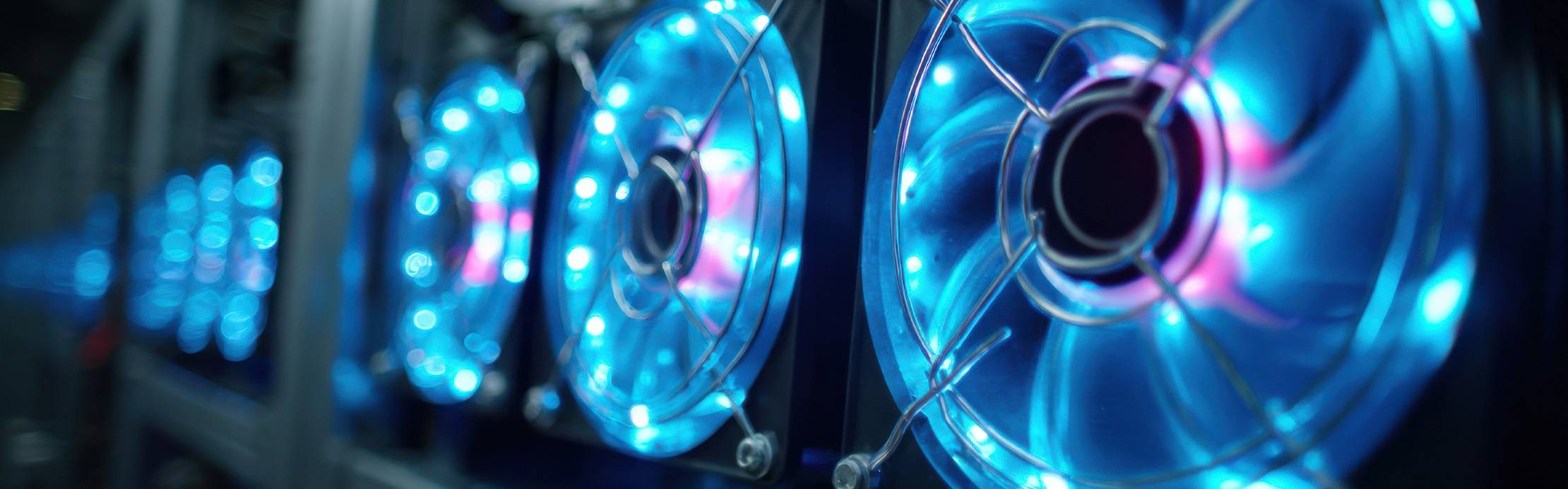 Nahaufnahme einer Reihe von Computerlüftern, die mit hellen blauen LEDs beleuchtet sind. Der vorderste Lüfter mit seinem runden Metallschutzgitter ist im Fokus, während sich die weiteren Lüfter in einer Fluchtlinie nach hinten reihen und zunehmend unscharf in einem Bokeh-Effekt verschwimmen. Das Bild erinnert an das Innere eines modernen Server-Racks oder eines leistungsstarken PC-Systems in dunkler Umgebung.