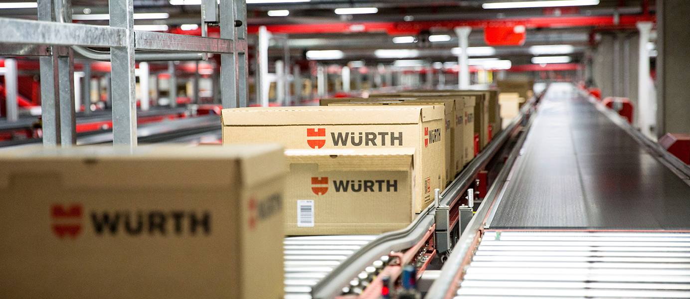  Dieses Teaserbild zeigt eine Reihe von braunen Würth-Kartons, die auf einem Fließband in einer großen Lagerhalle transportiert werden, mit weiteren Fließbändern und roten Metallstrukturen im Hintergrund.