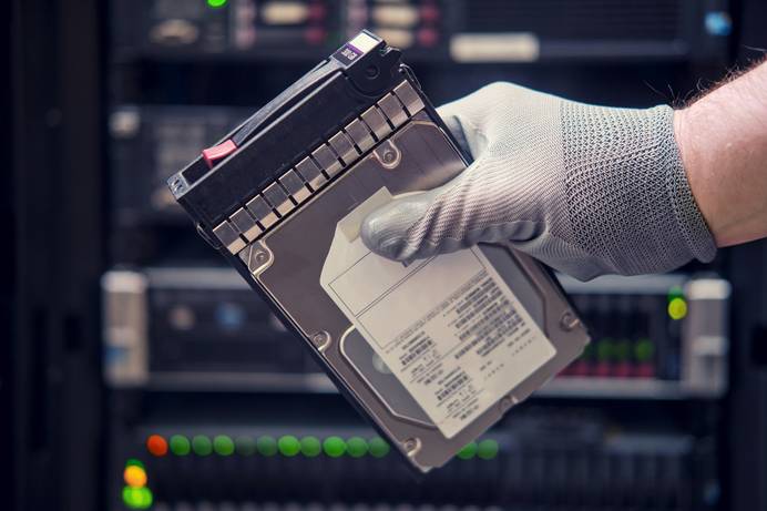 Jemand hält eine Festplatte in der Hand. Die Person trägt einen Handschuh. Im Hintergrund ist ein Server-Rack zu sehen.