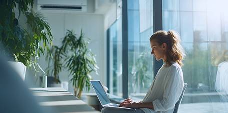 Eine Frau arbeitet an einem Laptop in einem hellen Büro mit Pflanzen. Mit Weiterleitung zur Seite  Sygonix Kühle Räume im Sommer
