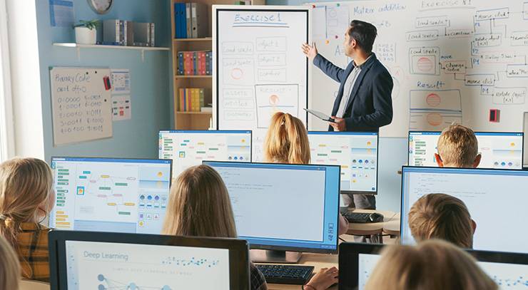 Lehrer unterrichtet Schüler an Computern in einem Informatikraum mit mehreren Monitoren.