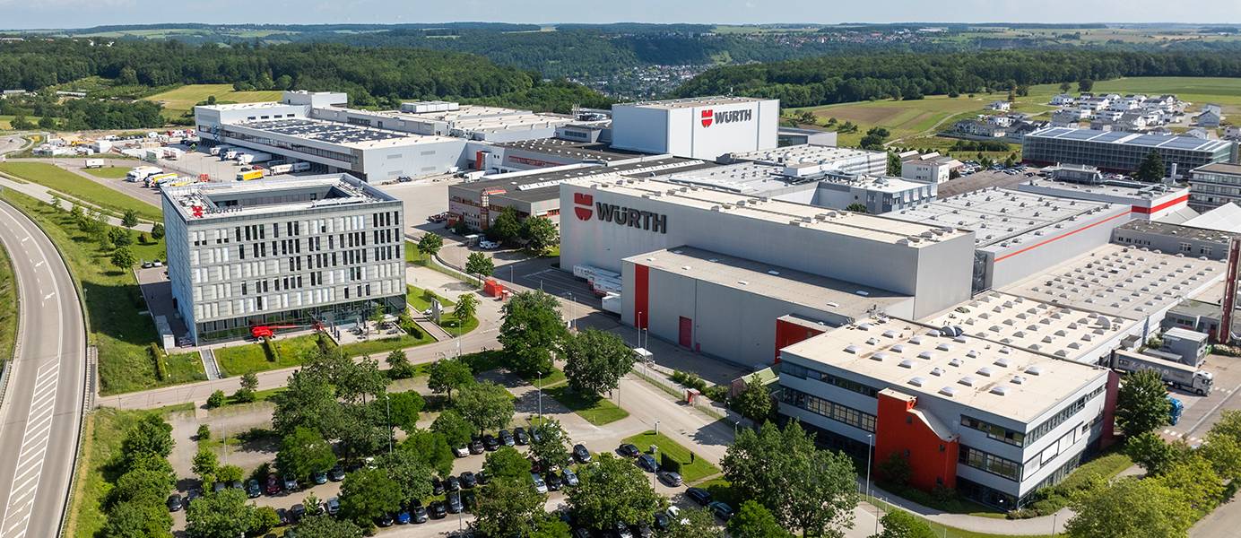  Dieses Teaserbild zeigt eine Luftaufnahme des Würth-Hauptsitzes, bestehend aus mehreren großen, modernen Gebäuden, umgeben von grünen Flächen, Parkplätzen und einer Straße, unter einem klaren blauen Himmel mit einigen weißen Wolken.