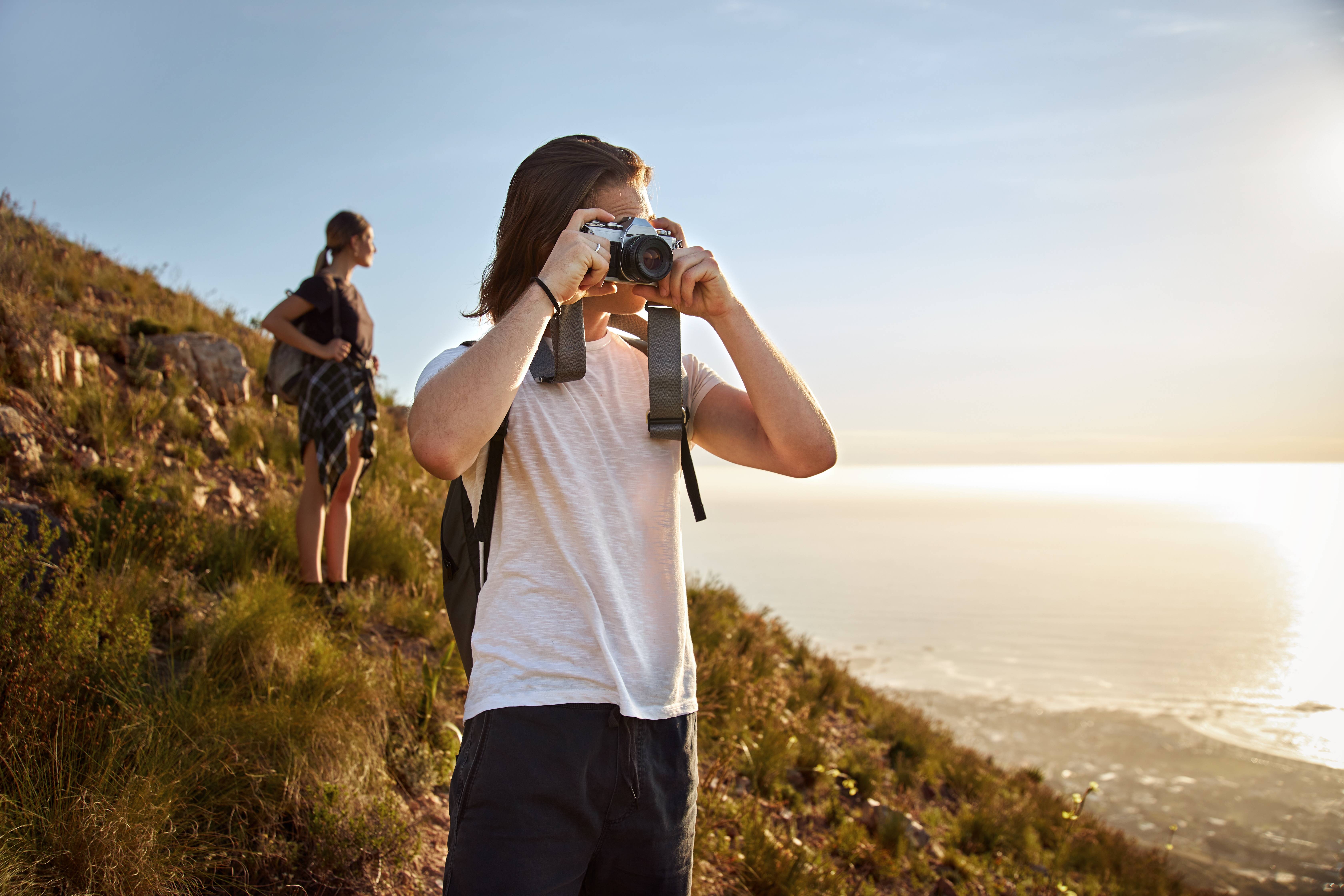 Ein Mann und eine Frau wandern durch die Natur. Sie stehen auf einem Hügel, im Hintergrund ist das Meer zu sehen. Der Mann macht mit einer Kamera ein Foto, die Frau steht hinter ihm und blickt Richtung Wasser.