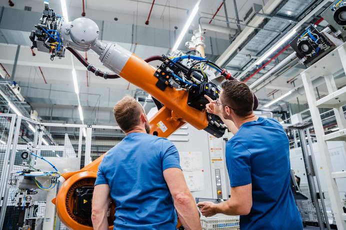 Zwei Männer in blauen T-Shirts stehen vor einem überdimensional großen Roboterarm mit orangefarbenem Korpus und blicken auf die komplexe, außen angebrachte Elektronik. Die Umgebung ist industriell geprägt. Im Hintergrund sind Trenngitter und eine Robotersteuerung in Form eines Schaltschranks zu sehen. An den Decken befinden sich helle Leuchtröhren.