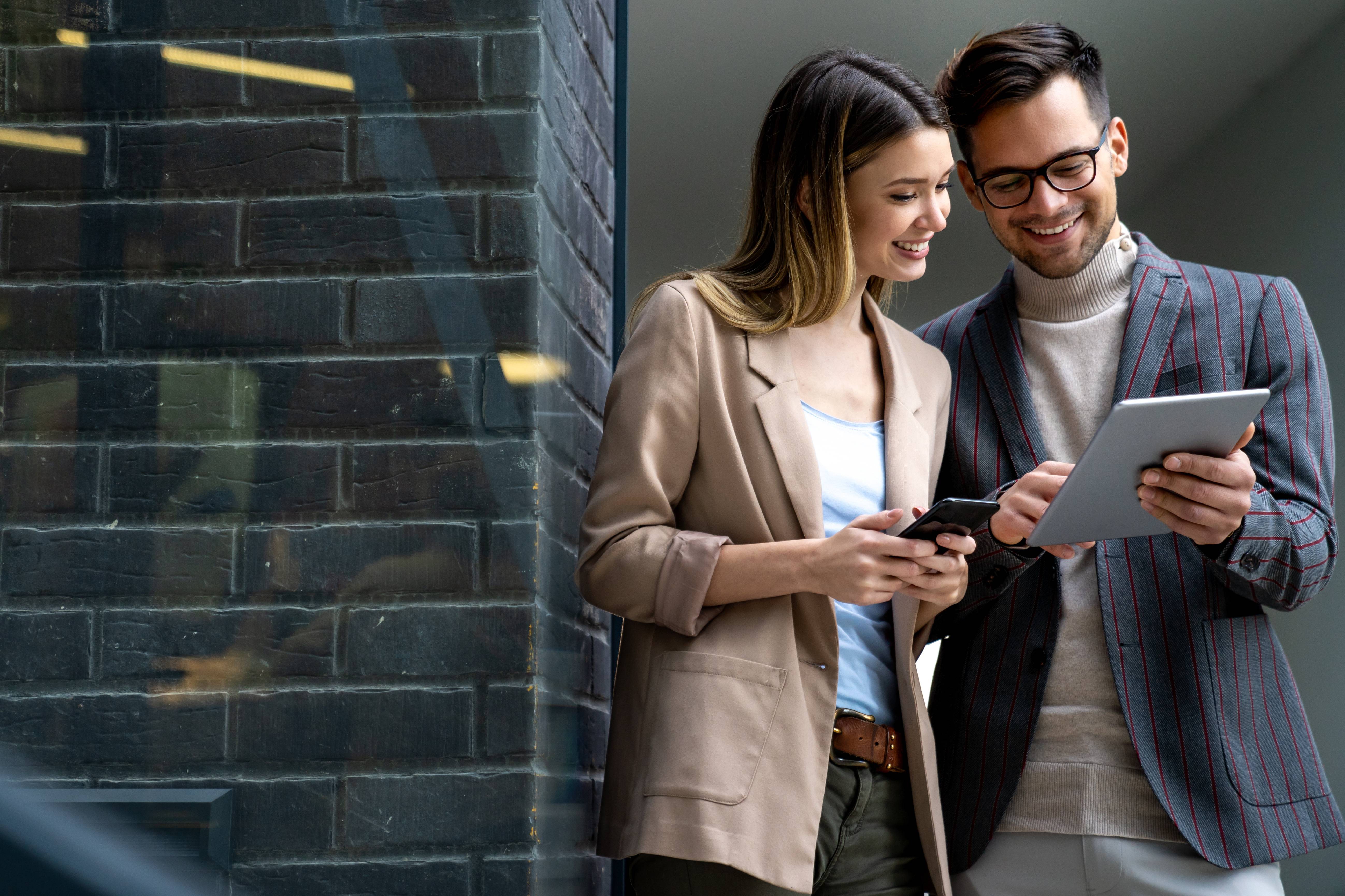Ein Mann und eine Frau stehen neben einer grau verklinkerten Mauer und schauen gemeinsam auf ein Tablet. Die Frau trägt einen hellbraunen Blazer, ein weißes Shirt und eine dunkle Hose. Sie hält ein Smartphone in der Hand. Der Mann trägt ein gestreiftes Sakko, einen Rollkragenpullover und eine helle Hose. Er hält das Tablet und zeigt mit der Hand auf das Display.