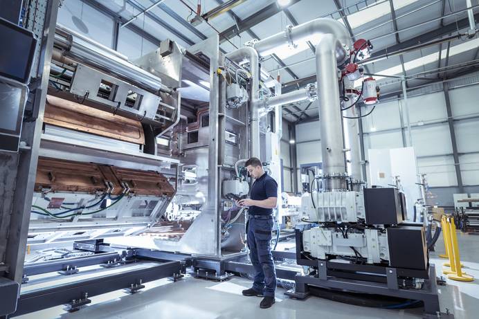 Ein Mann in Arbeitskleidung steht seitlich vor einer großen Vakuumbeschichtungsanlage mit dicken Rohren und technischen Apparaturen aus Edelstahl. Er schaut auf ein Tablet, das er in den Händen hält.