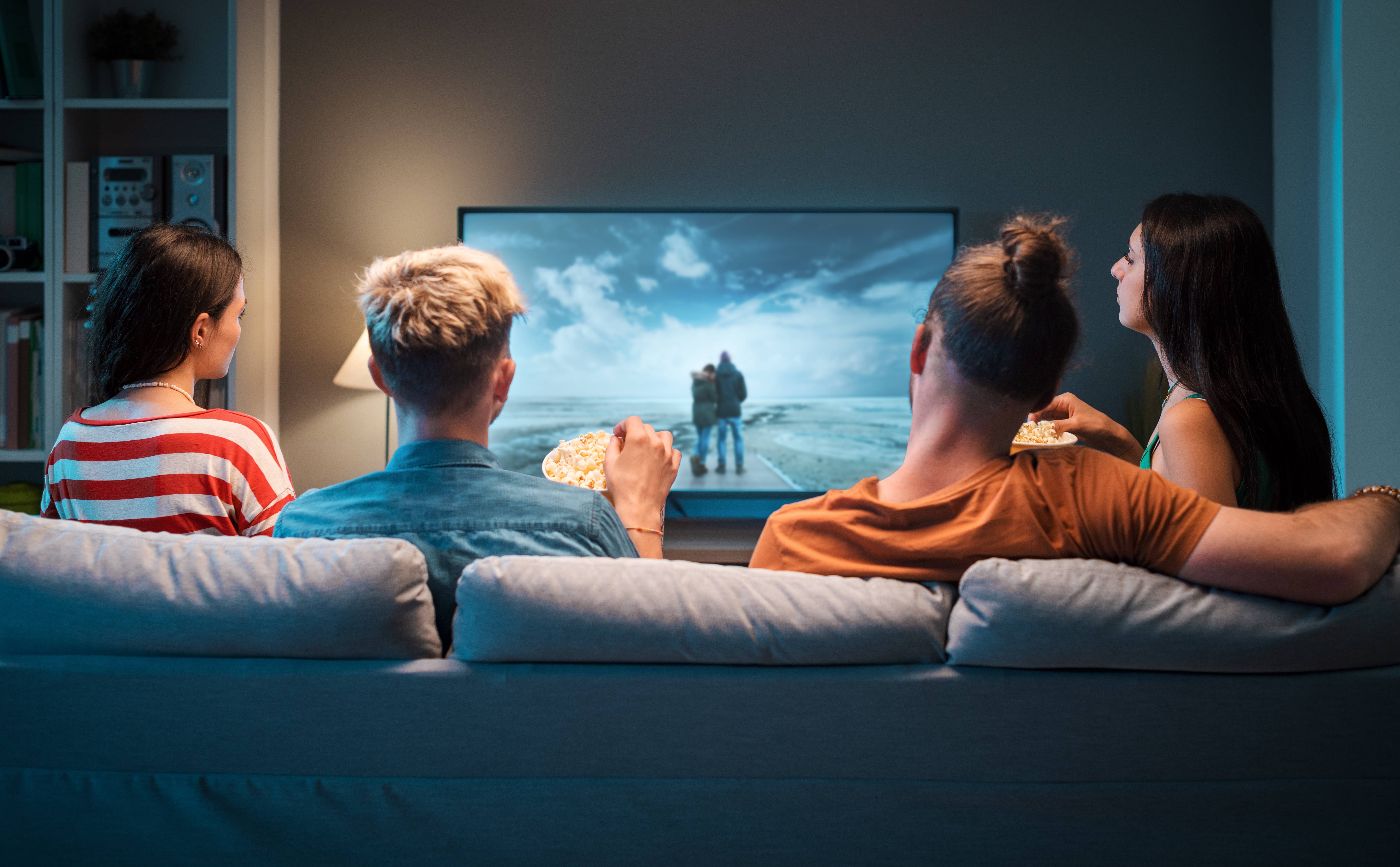 Vier Personen sitzen auf einem Sofa, essen Popcorn und schauen gemeinsam einen Film auf einem großen Fernseher.