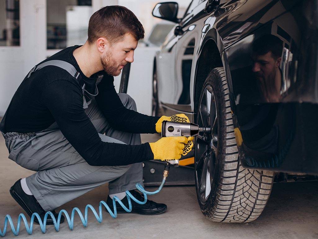 Ein Mann mit schwarzem, langärmeligem Shirt und grauer Arbeitshose kniet vor einem schwarzen Fahrzeug. An dessen Hinterreifen hält er einen druckluftbetriebenen Schraubendreher, von dem ein blauer Spiralschlauch abgeht.