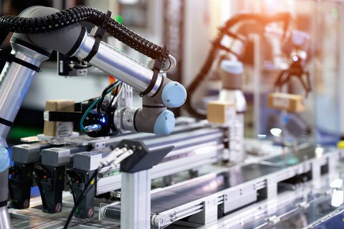 Ein Roboterarm mit komplexer Elektronik als Teil in einer Produktionsanlage. Vor ihm befindet sich ein Förderband. Im Hintergrund ist ein Roboter mit Greifer zu sehen, der verpackte Ware vom Band hebt.