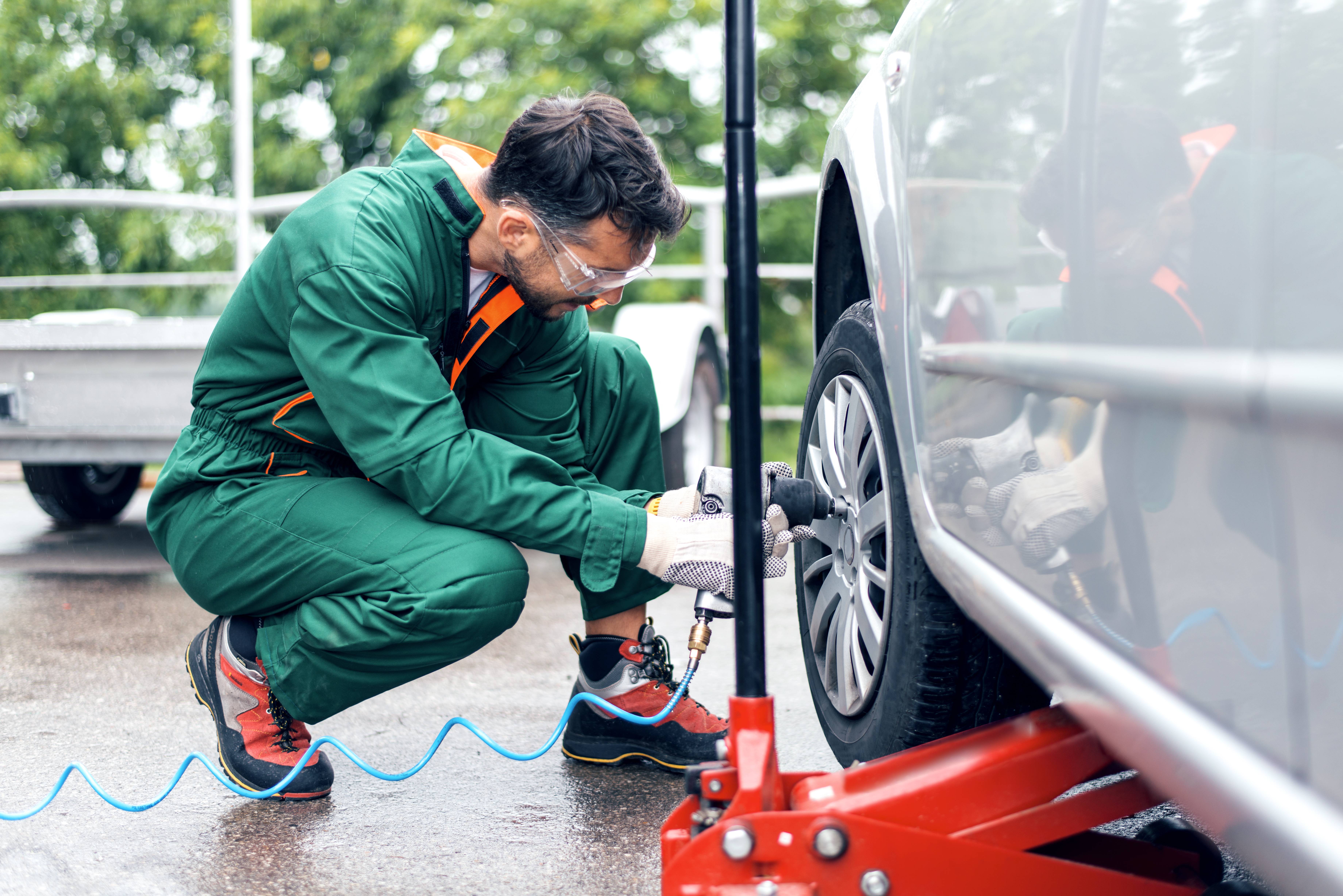 Ein Mann mit grünem Overall, Arbeitsschuhen und Schutzbrille kniet vor einem silberfarbenen Auto und löst die Schrauben am Vorderrad.