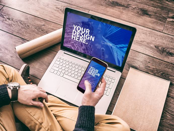 Person sitzt auf Holzboden und nutzt einen Laptop, während sie ein Smartphone in der Hand hält.