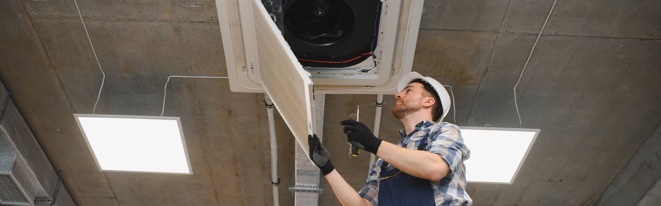 Untersicht eines Technikers mit weißem Schutzhelm und Arbeitshandschuhen, der ein an einer Betondecke montiertes Klimagerät wartet. Er hat die Filterabdeckung der Anlage heruntergeklappt, hält sie mit einer Hand fest und blickt mit einem Schraubenzieher in der anderen Hand konzentriert in das Innere des Geräts.