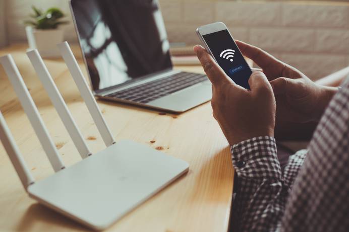 Ein Mann sitzt an seinem Arbeitsplatz. Auf dem Tisch stehen ein Laptop und ein WLAN-Router. Der Mann hält ein Smartphone in den Händen. Auf dessen Bildschirm ist zu sehen, dass es gerade Verbindung zum WLAN aufnimmt.