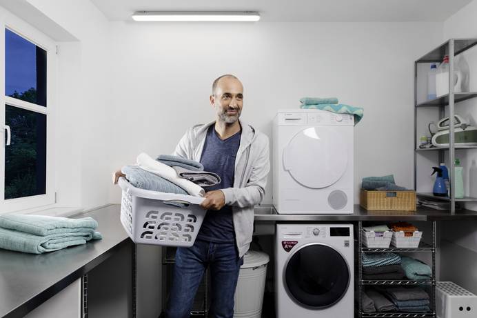 Ein leger gekleideter Mann steht in einem funktional eingerichteten Wirtschaftsraum und trägt einen mit Handtüchern gefüllten Wäschekorb. Hinter ihm befinden sich ein Wäschetrockner und eine Waschmaschine. Die Waschmaschine steht unter einer Arbeitsplatte aus Edelstahl. Rechts sind Regale mit Aufbewahrungskörben und weiteren Handtüchern zu sehen. Links befindet sich ein Fenster, das den Blick auf den dämmernden Himmel freigibt.