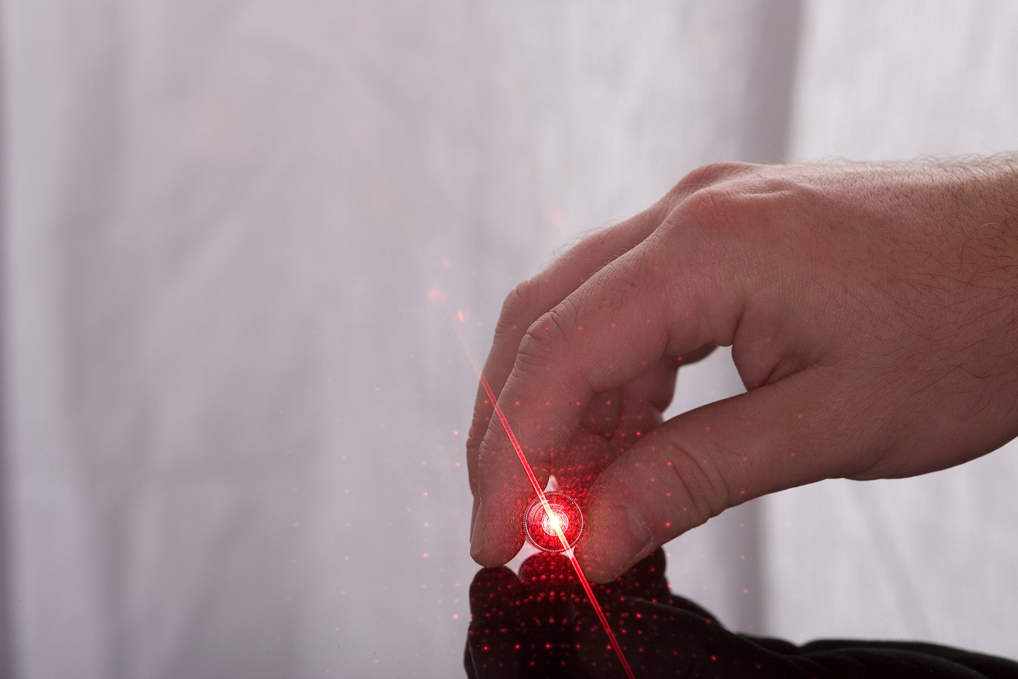 Das Bild zeigt eine Hand, welche einen kleinen, runden Laserpointer zwischen Daumen und Zeigefinger hält. Aus dem Laserpointer tritt ein leuchtend roter Laserstrahl aus. 