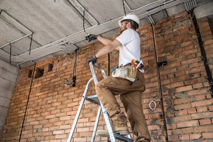 Mann mit Arbeitsschutz steht auf einer Leiter vor einer gemauerten Wand, aus der mehrere Leitungen und Kabel herausragen.