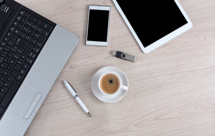 Man sieht einen Schreibtisch von oben, auf dem ein Laptop, ein Kugelschreiber, en Smartphone, ein Tablet, eine Tasse Kaffee und ein USB-Stick liegen.
