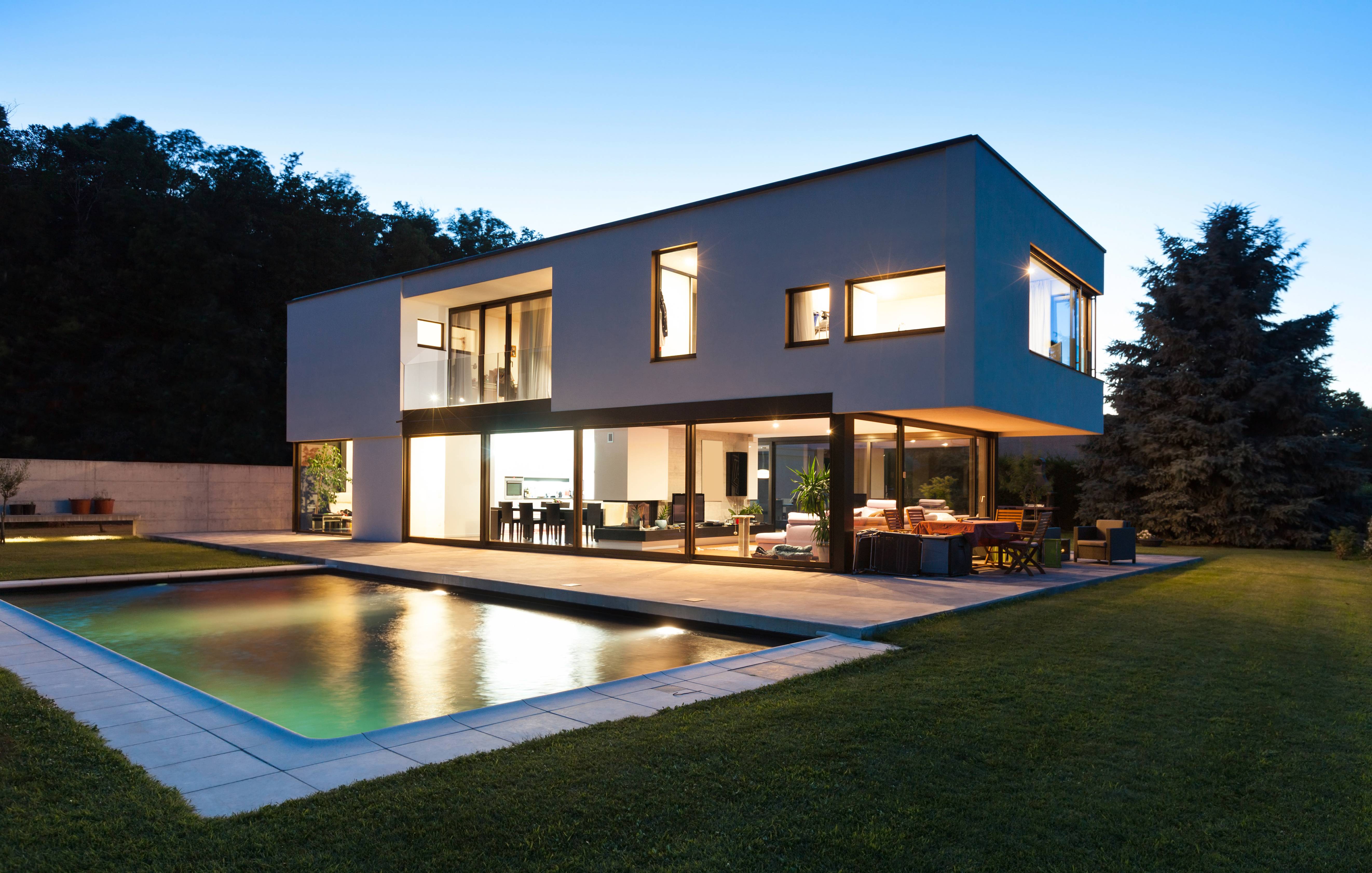 Moderna, dvonadstropna hiša z velikimi okni in bazenom v ospredju, mrak.