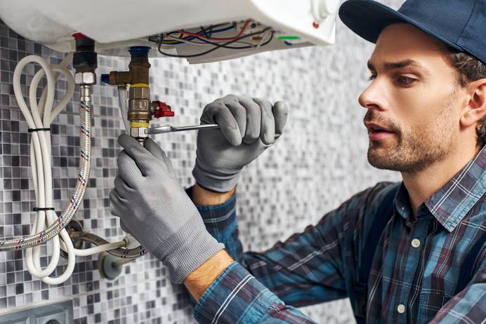 Das Bild zeigt einen Installateur oder Handwerker bei der Arbeit an einer Wasser- oder Heizungsanlage. Er trägt eine blaue Kappe, ein kariertes Hemd und graue Arbeitshandschuhe. Mit einem Schraubenschlüssel zieht er eine Verbindung an einem Rohrsystem unter einem Boiler oder einer Heizung fest. Die Umgebung ist sauber und modern, mit einer Wand aus kleinen Mosaikfliesen im Hintergrund. Kabel und Schläuche sind ordentlich befestigt, was auf eine professionelle Installation hinweist. Die konzentrierte Miene des Handwerkers deutet darauf hin, dass er mit Präzision an der Reparatur oder Wartung arbeitet.