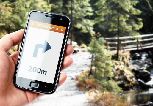Ein Handy mit einer Wegbeschreibung (in 200m links abbiegen). In Hintergrund ist ein Waldweg mit Bachlauf zu sehen.