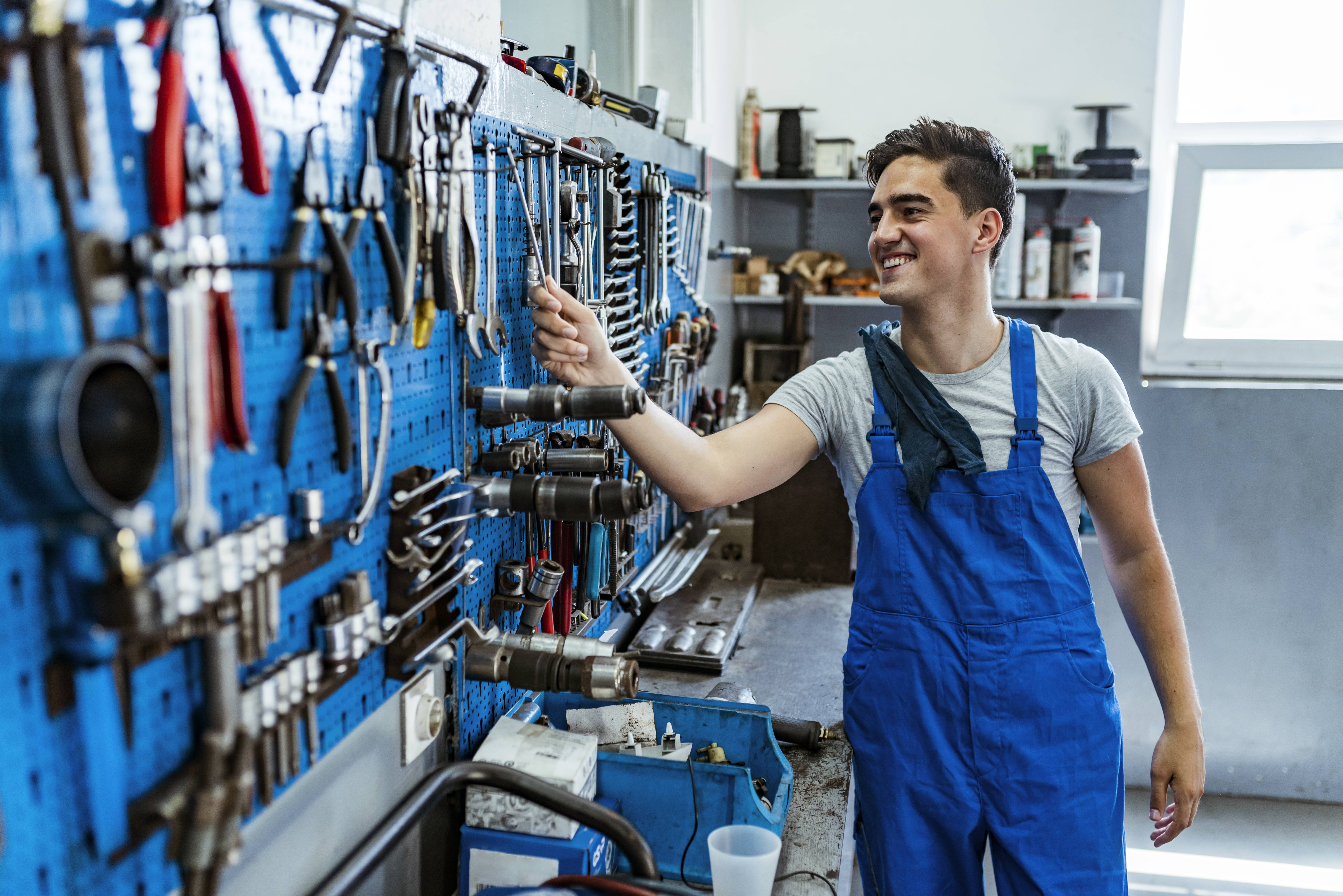 Lächelnder Handwerker in blauer Latzhose greift in einer Werkstatt nach einem Schraubenschlüssel von der Werkzeugwand.