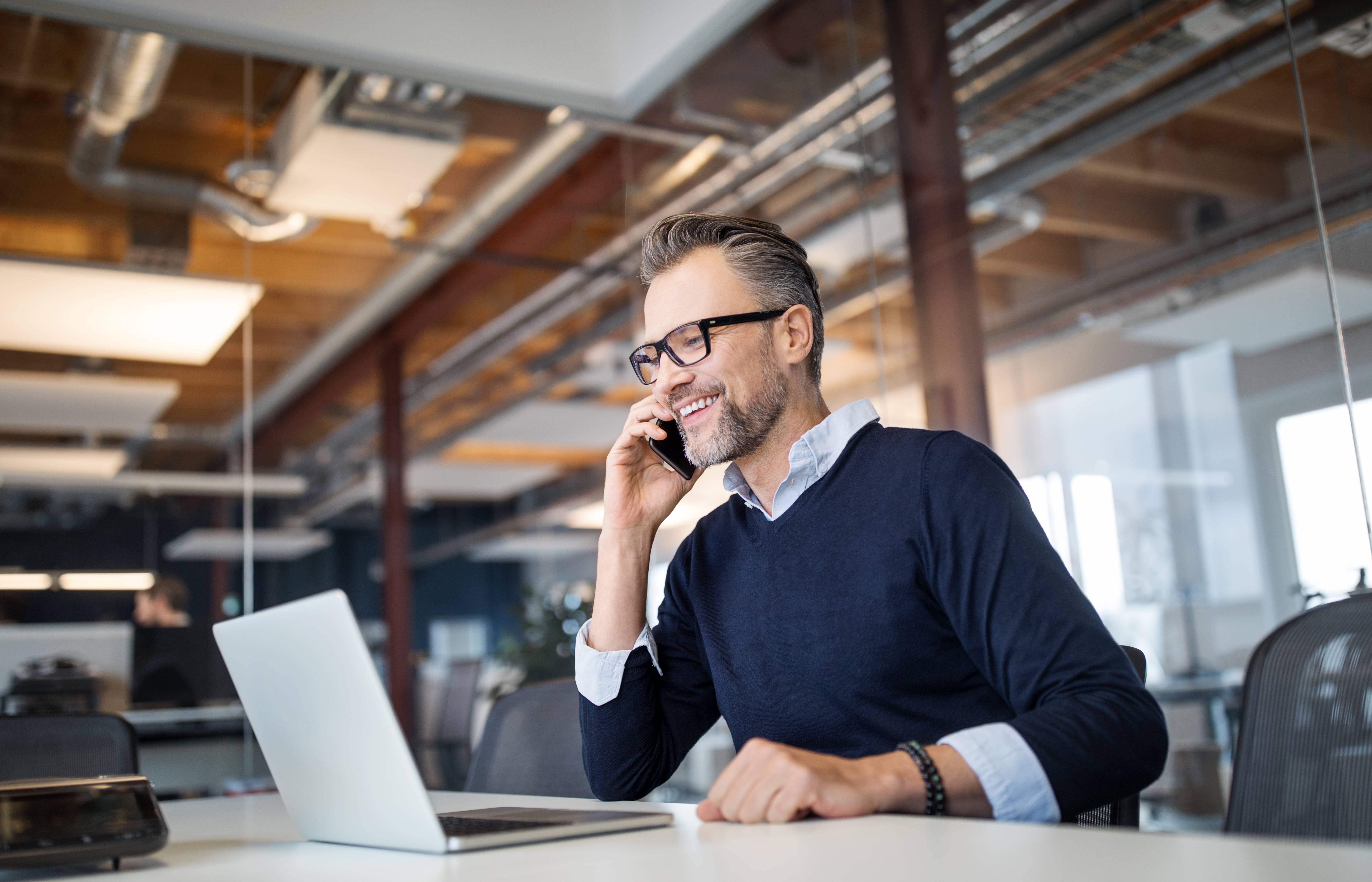 Un homme portant des lunettes téléphone en souriant au bureau tout en travaillant sur un ordinateur portable.