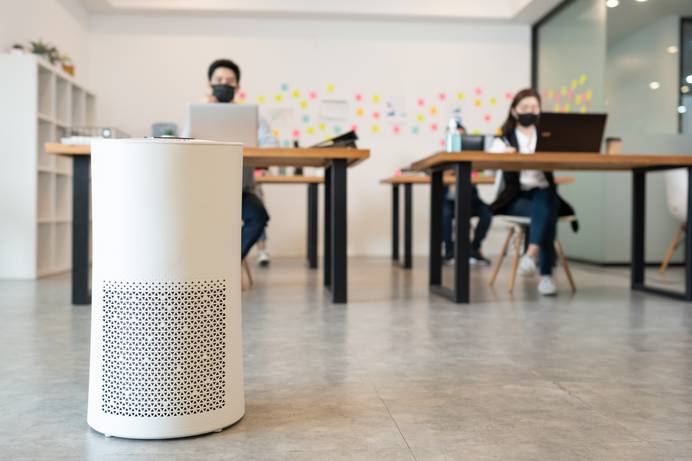 Modernes Büro mit zwei Schreibtischen im Hintergrund, an denen jeweils ein Mensch mit Maske sitzt. Im Vordergrund steht ein Luftreiniger auf dem Boden.