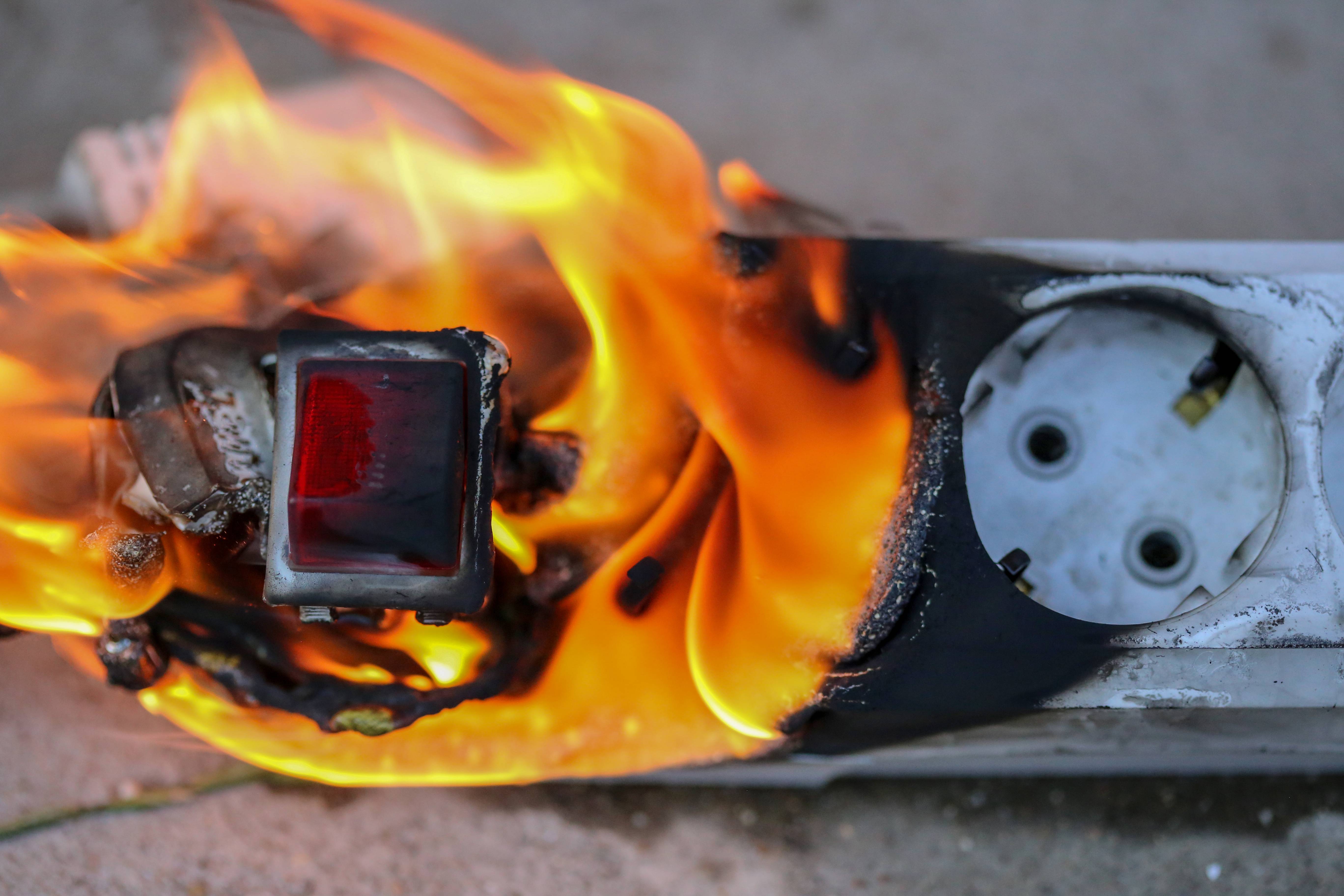 Großaufnahme einer weißen Verteilerdose mit An-/Aus-Schalter, die auf einem steinernden Untergrund liegt. Weite Teile der Dose sind verschmort und stehen in Flammen.