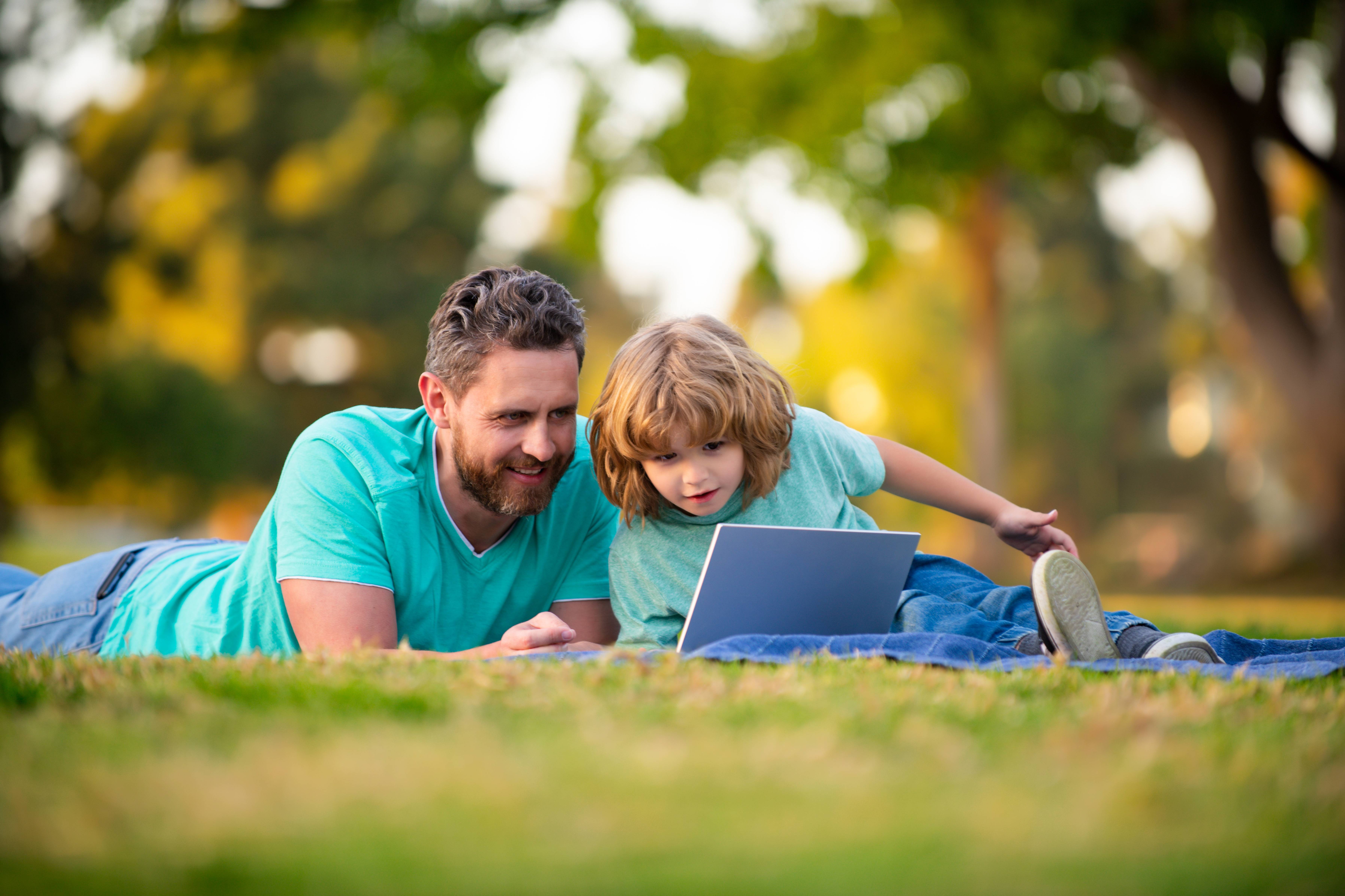 Vater und Sohn liegen auf einer Rasenfläche im Park. Sie tragen beide ein türkises T-Shirt und eine blaue Jeans und schauen gemeinsam auf den Bildschirm eines Tablets. Rundherum ist viel Grün zu sehen.