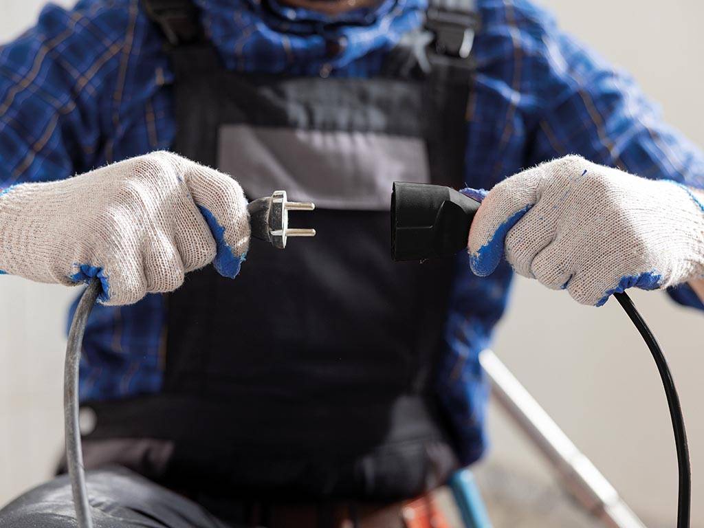 Ein Mann mit kariertem Hemd und Arbeitshose hält einen Stecker und eine Buchse vor sich und ist im Begriff, beides zusammenzustecken.