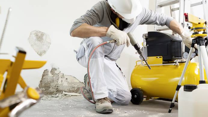 Ein Handwerker mit Helm hält in der rechten Hand eine Luftdruckpistole. Neben ihm befindet sich ein gelber Drucklufttank mit zwei analogen Skalen. Der Mann beugt sich seitlich darüber, um Druckeinstellungen vorzunehmen. Im Hintergrund ist eine Wand mit Stellen von Mauerwerk und Putz zu sehen. Rechts vorne stehen ein Stativ und ein Kanister.