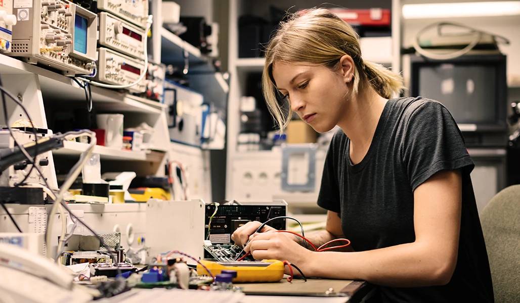 Eine junge Frau arbeitet konzentriert an elektronischen Geräten in einem Labor, umgeben von Technik. Das Bild ist in Blautönen gehalten.