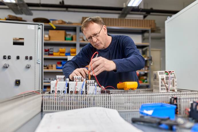 Ein Elektriker vor einer offenen Schalttafel in Werkstattumgebung hält die Prüfspitze einer roten Messleitung an ein Bauteil.