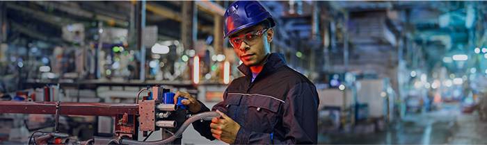 Uomo con cappello rigido blu e occhiali di sicurezza, in tenuta da lavoro, che lavora su macchinari in un ambiente industriale.