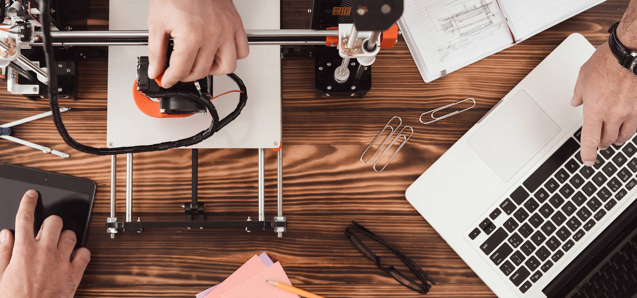 Draufsicht auf einen Arbeitsplatz mit 3D-Drucker, Laptop, technischen Skizzen und Arbeitsmaterialien für den Prototypenbau.