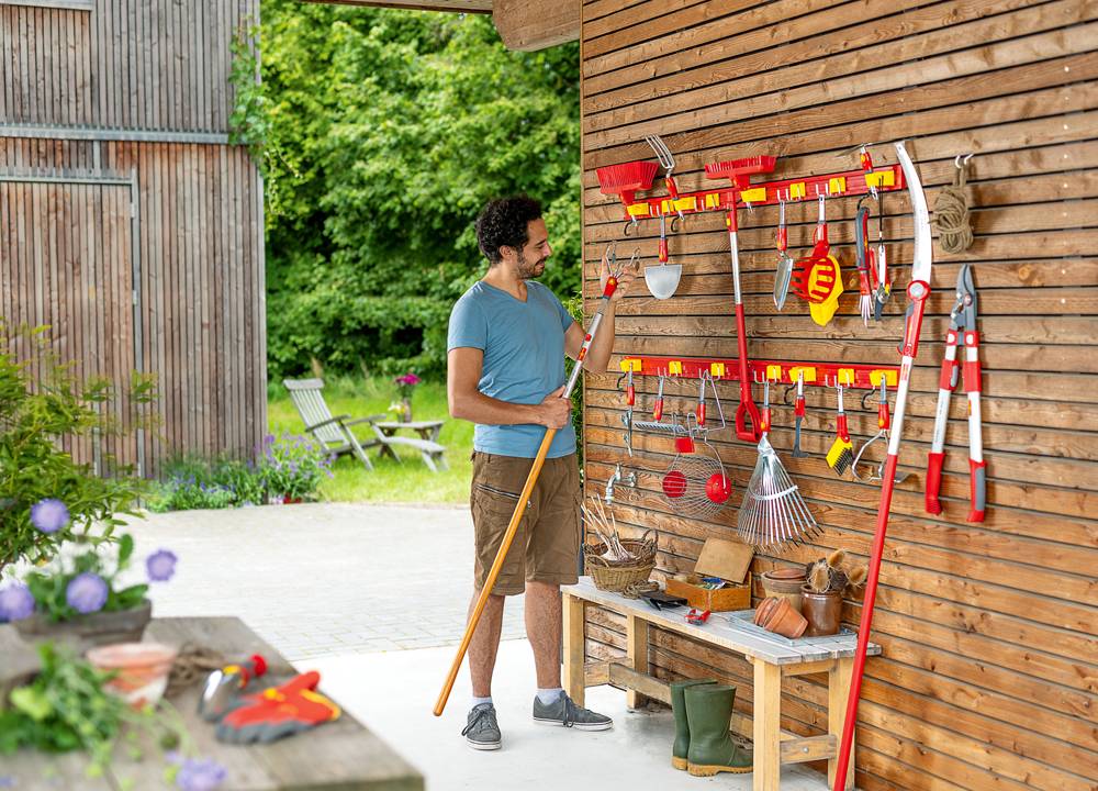 Mann hält Rechen in Garten mit Holzschuppen. Rote Gartengeräte an Wand montiert. Draußen Tisch, Stühle, grüne Pflanzen.