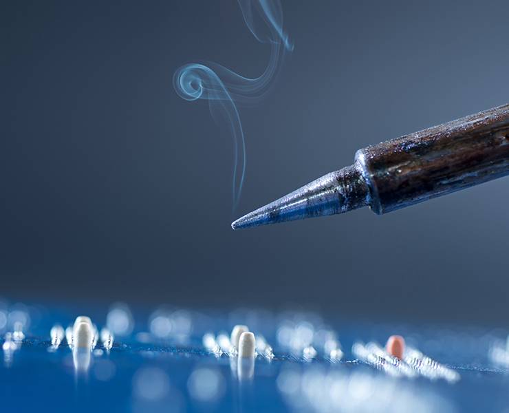 Über eine blaue Platine schwebt eine Lötkolbenspitze woraus Rauch entflieht