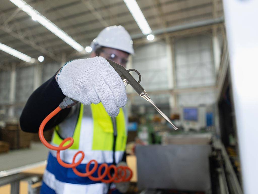 Mann in einer Industrieumgebung. Er trägt Schutzhelm, Schutzbrille, Schutzweste und Schutzhandschuhe. Die rechte Hand ist im Vordergrund zu sehen und hält eine Druckluftpistole, von der ein orangefarbener Spiralschlauch abgeht.
