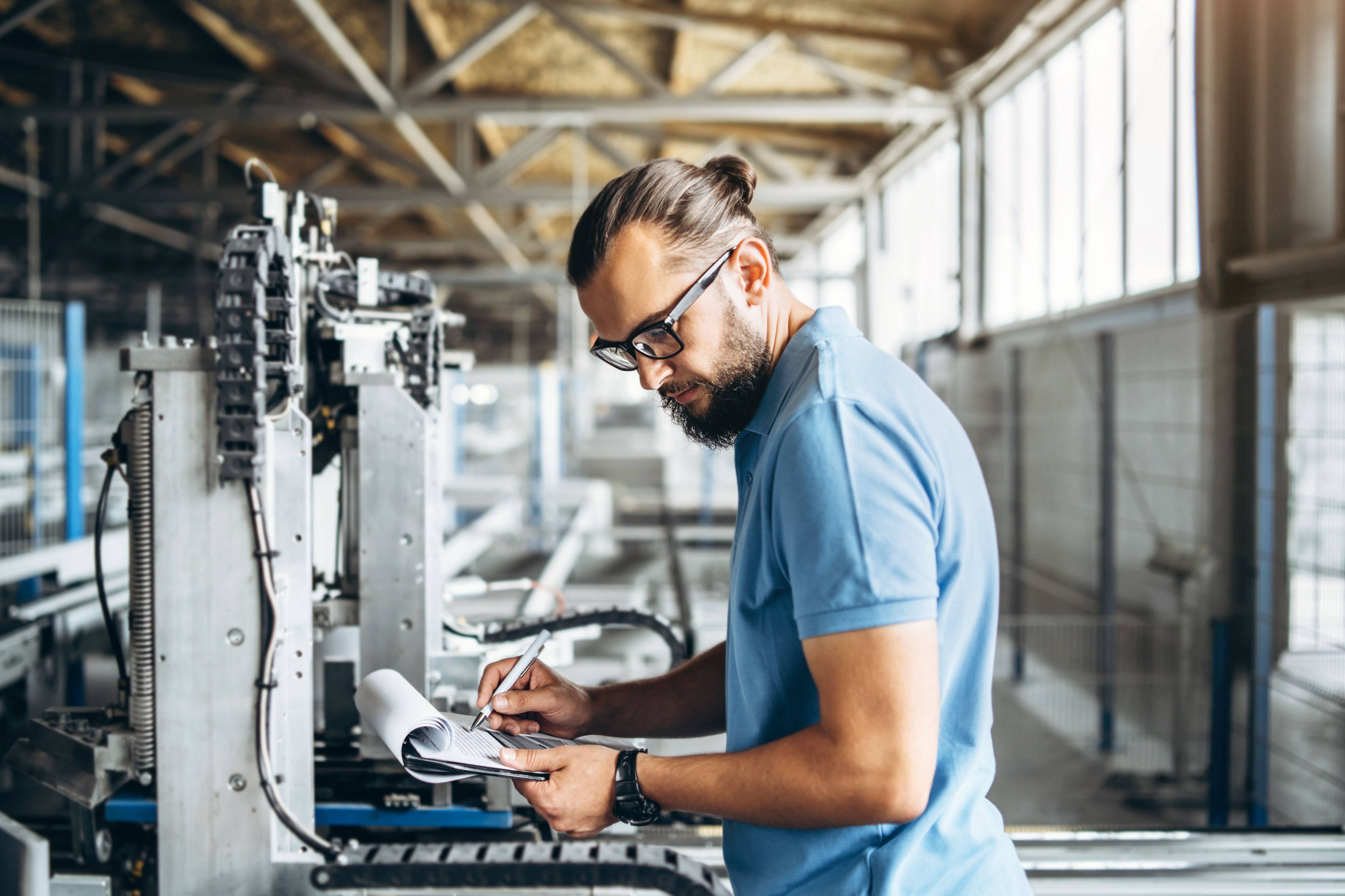 Un homme portant des lunettes prend des notes sur un bloc-notes devant une machine dans un atelier.