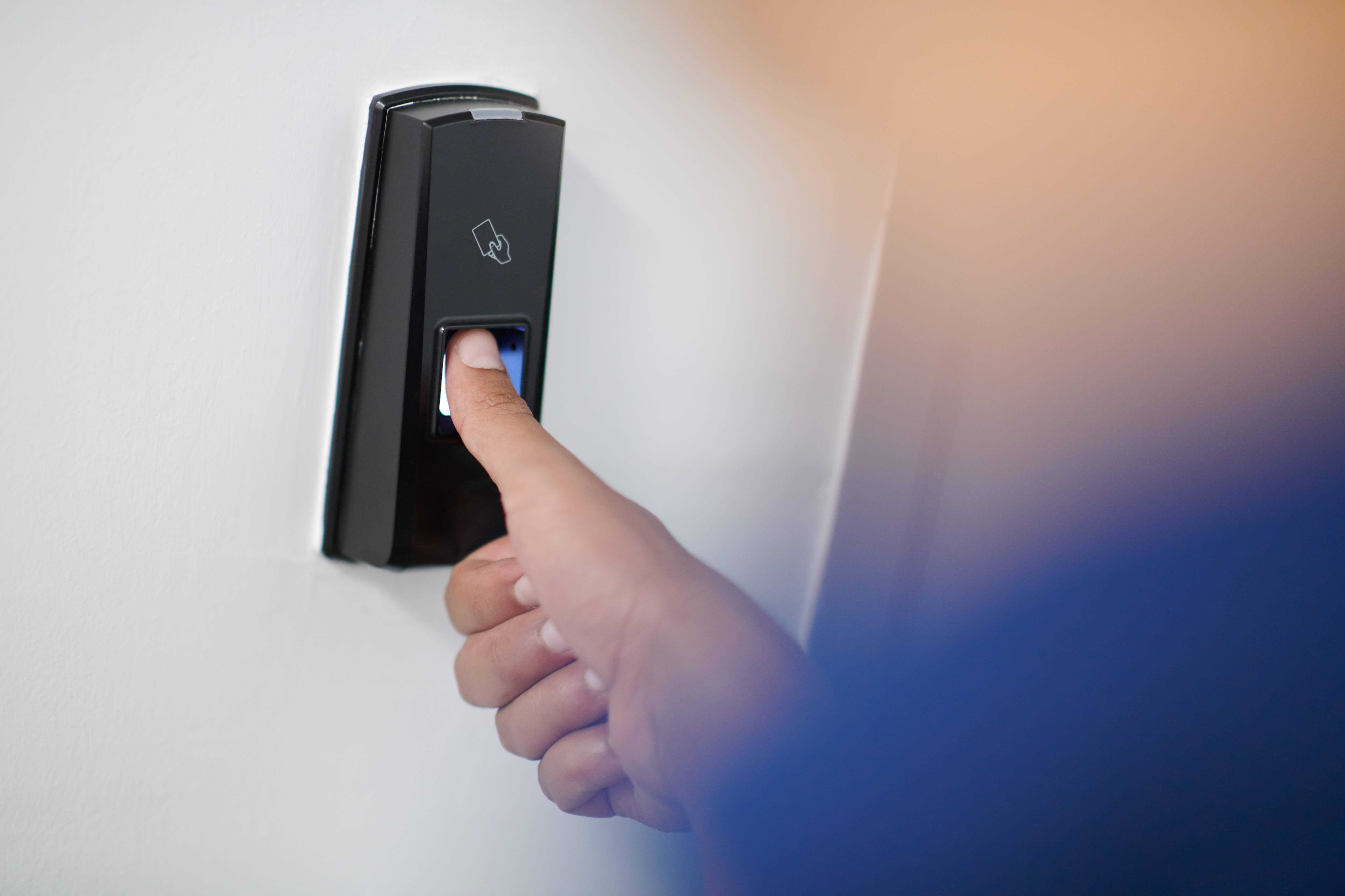 Ein schwarzer Fingerabdruckscanner an einer weißen Wand, der betätigt wird. Der Daumen wird gescannt. Im Vordergrund ist verschwommen eine Person zu erkennen.