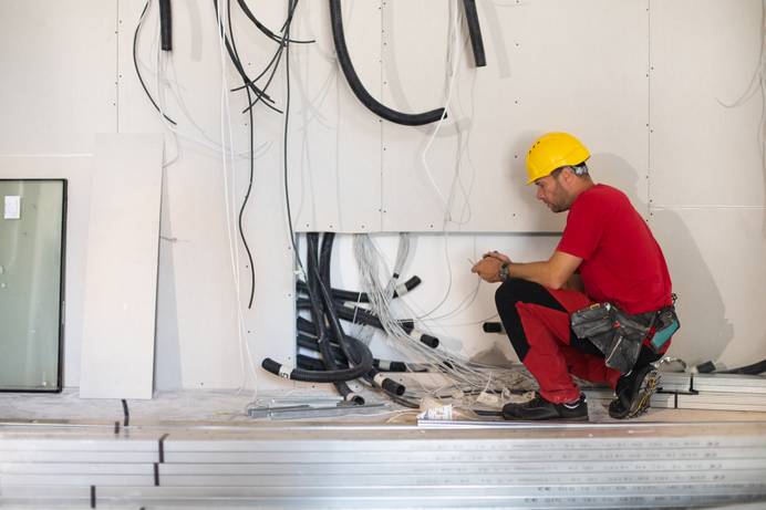 Diverse Kabel und Schläuche, die aus einer teilweise verkleideten Wand herausragen. Ein Mann mit Bauhelm kniet davor.