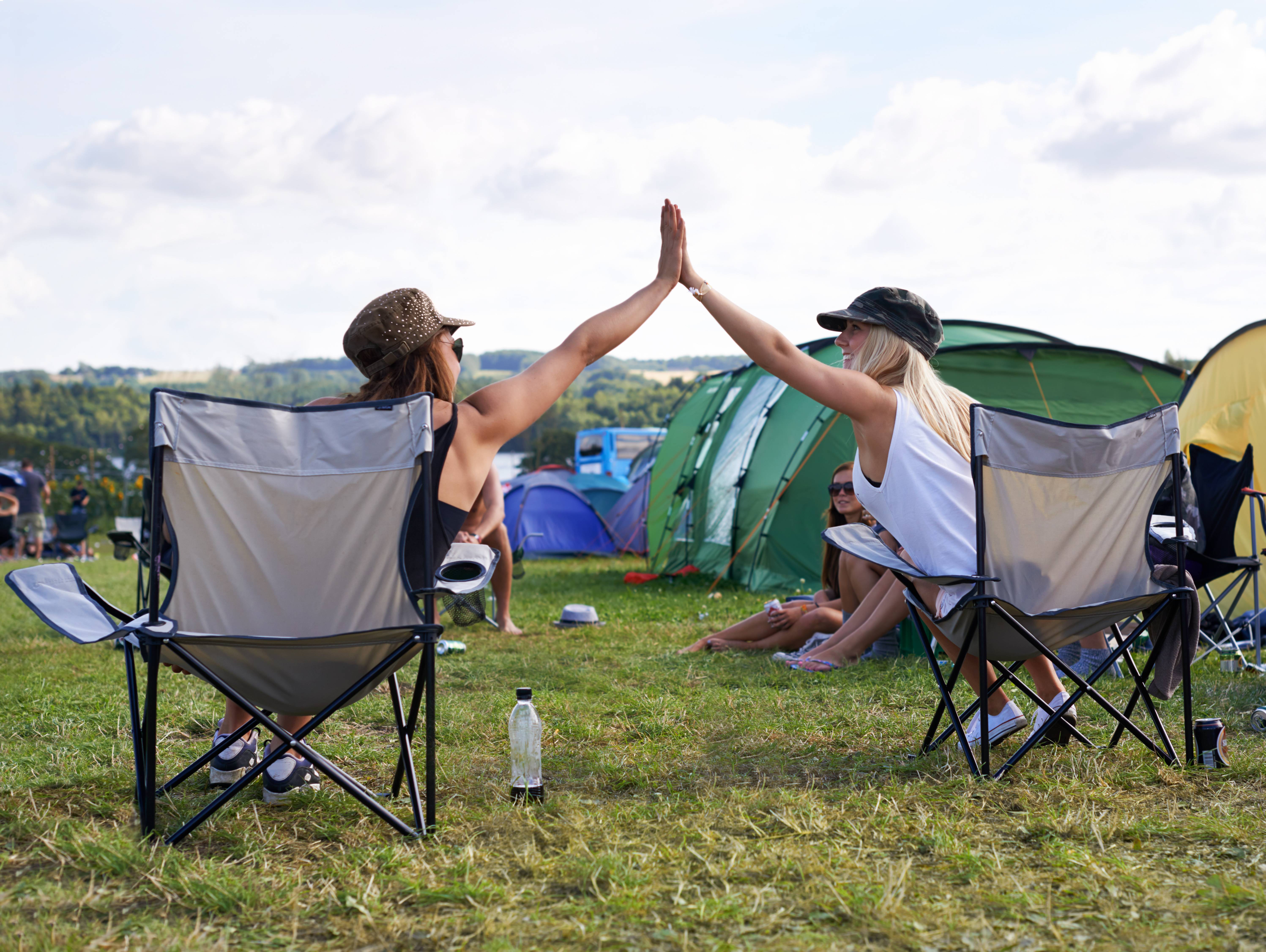 Deux femmes assises sur des chaises de camping devant des tentes se tapent dans la main sur le site d'un festival.