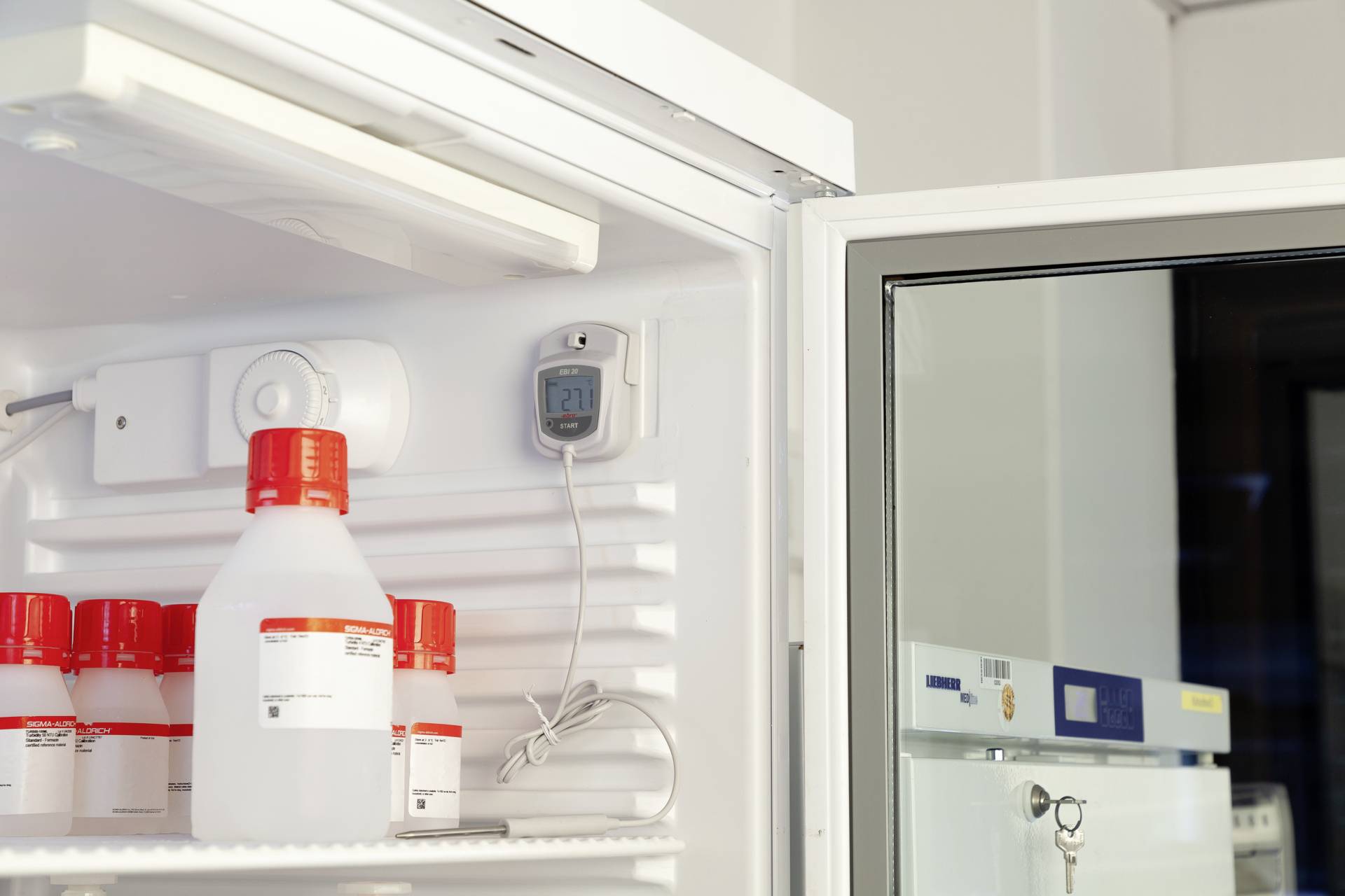 Otevřená lednice s plastovými lahvemi, některé jsou popsané. Teploměr ukazuje 21 stupňů Celsia. Okolí vypadá jako laboratoř.