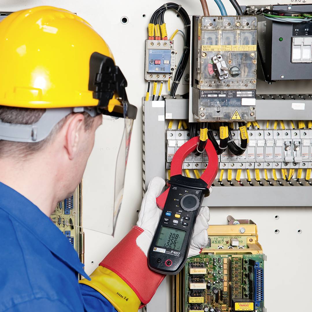 Osoba v bezpečnostním vybavení kontroluje elektrické komponenty multimetrem uvnitř rozvodné skříně.