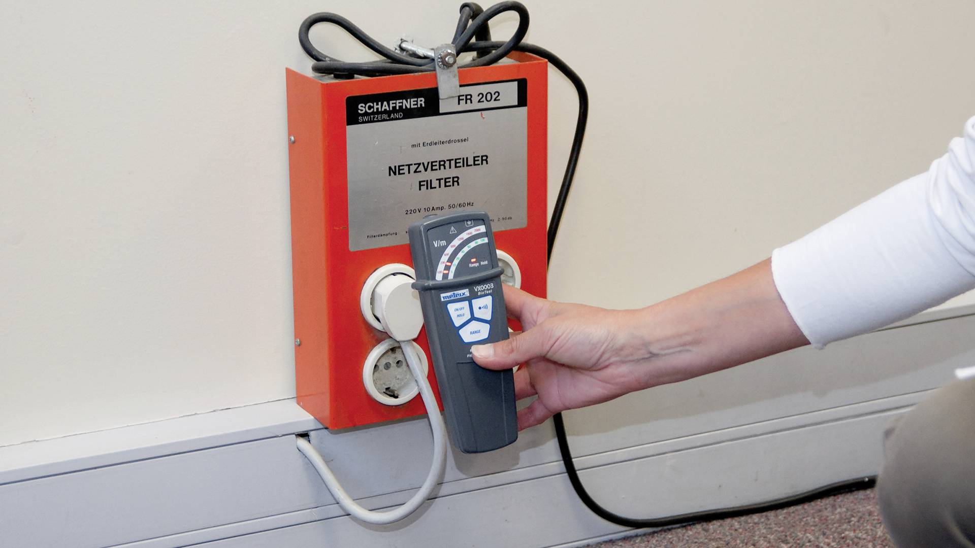 Osoba testuje zásuvku na červeném síťovém filtru na stěně pomocí měřicího přístroje. Měřicí přístroj zaznamenává elektrické hodnoty.