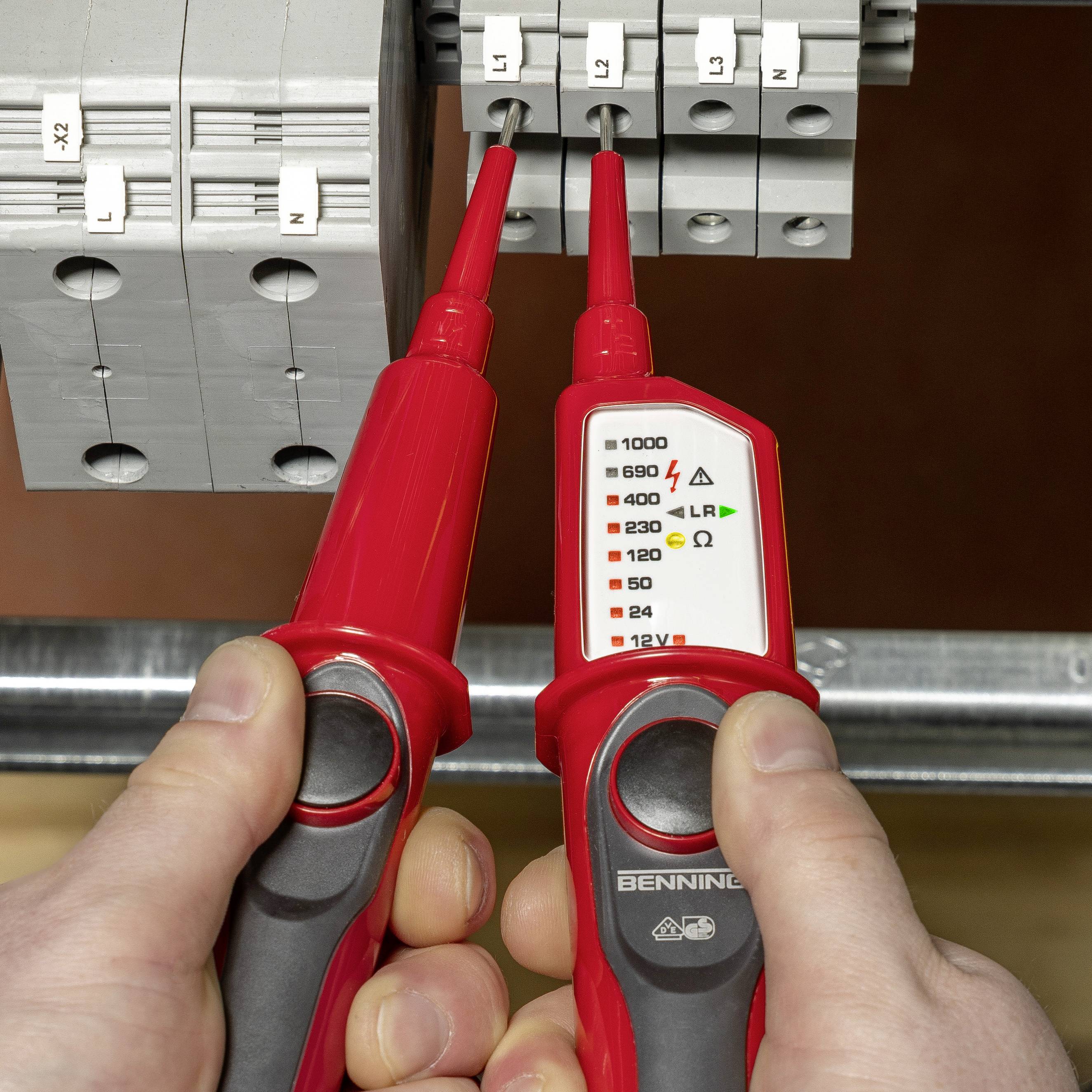 Osoba drží dva červené zkoušečky napětí před šedou elektrorozvodnou skříní, aby změřila elektrické napětí.